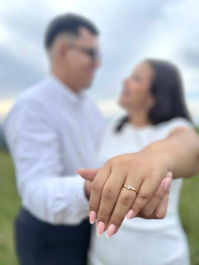 La mano de Jennyfer con el anillo de compromiso en primer plano, la pareja desenfocada detrás