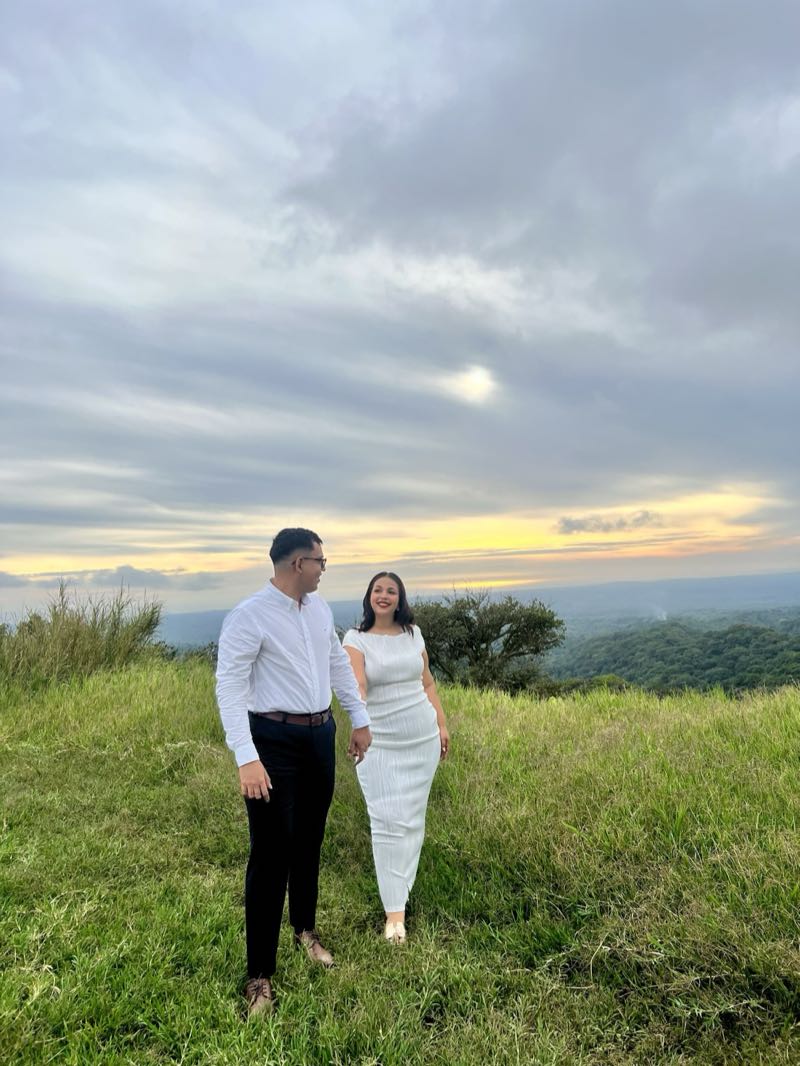 Byron y Jennyfer tomados de la mano en un mirador de pasto verde, al atardecer